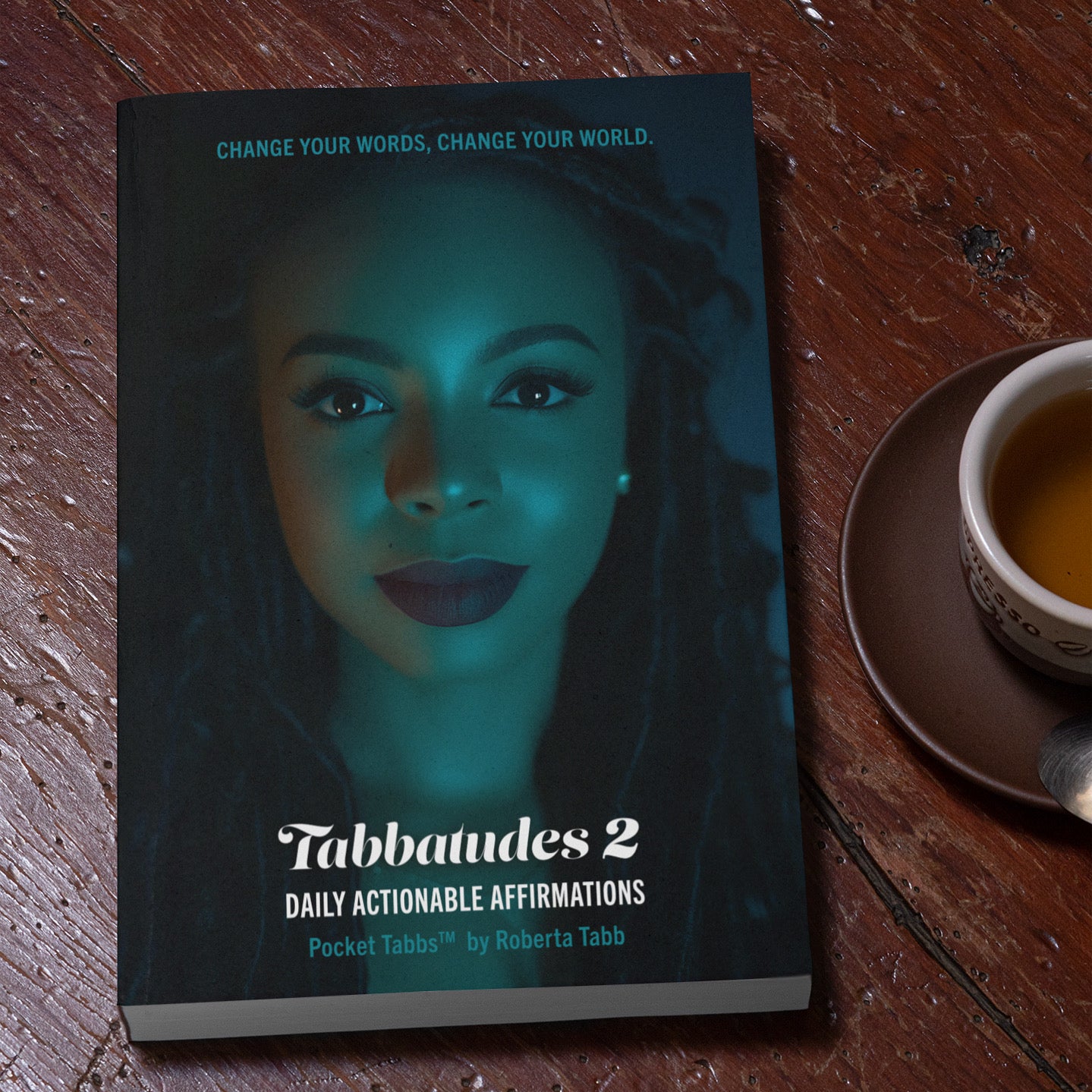 Book titled 'Tabbaudes 2' with a woman's face on the cover, placed on a wooden surface next to a cup of tea.