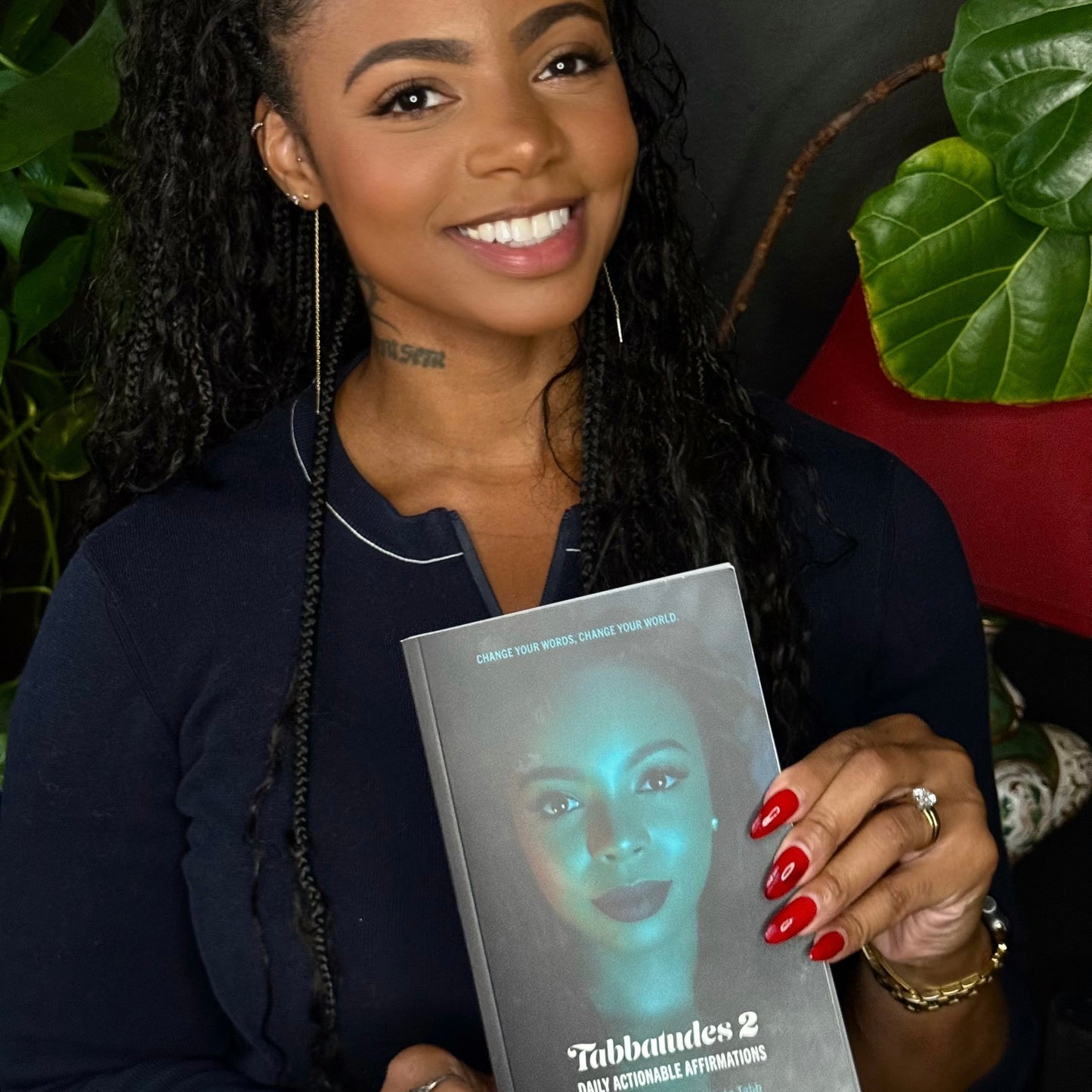 Roberta Tabb holding a book titled 'Tabbandes 2' with a blurred face on the cover, surrounded by greenery.