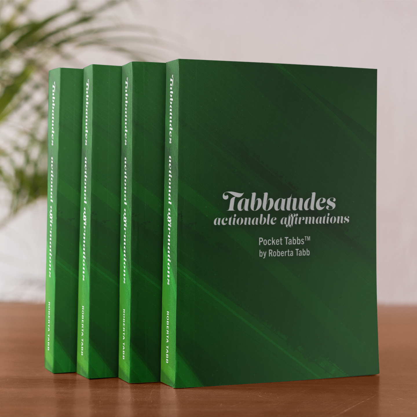 Four green books titled 'Tabbaudes actionable affirmations' on a wooden surface with a blurred plant in the background.