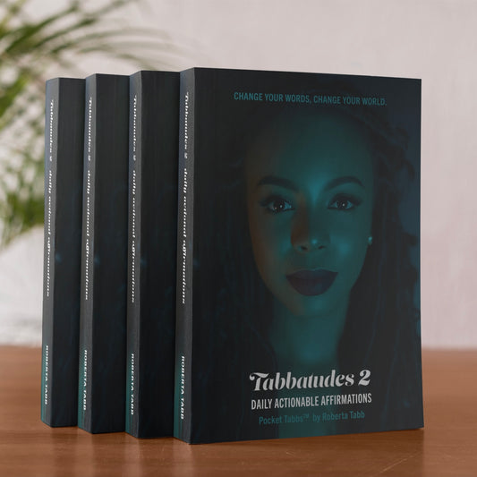 Four black books titled 'Tabbatudes 2' by Roberta Tabb on a wooden surface with a blurred plant in the background.